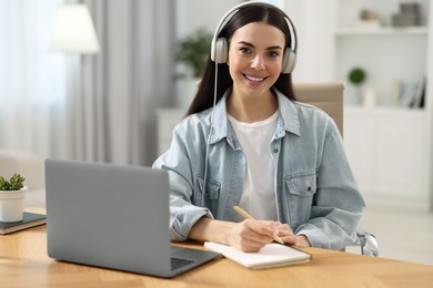 Photo of Young woman in headphones watching webinar at table in room