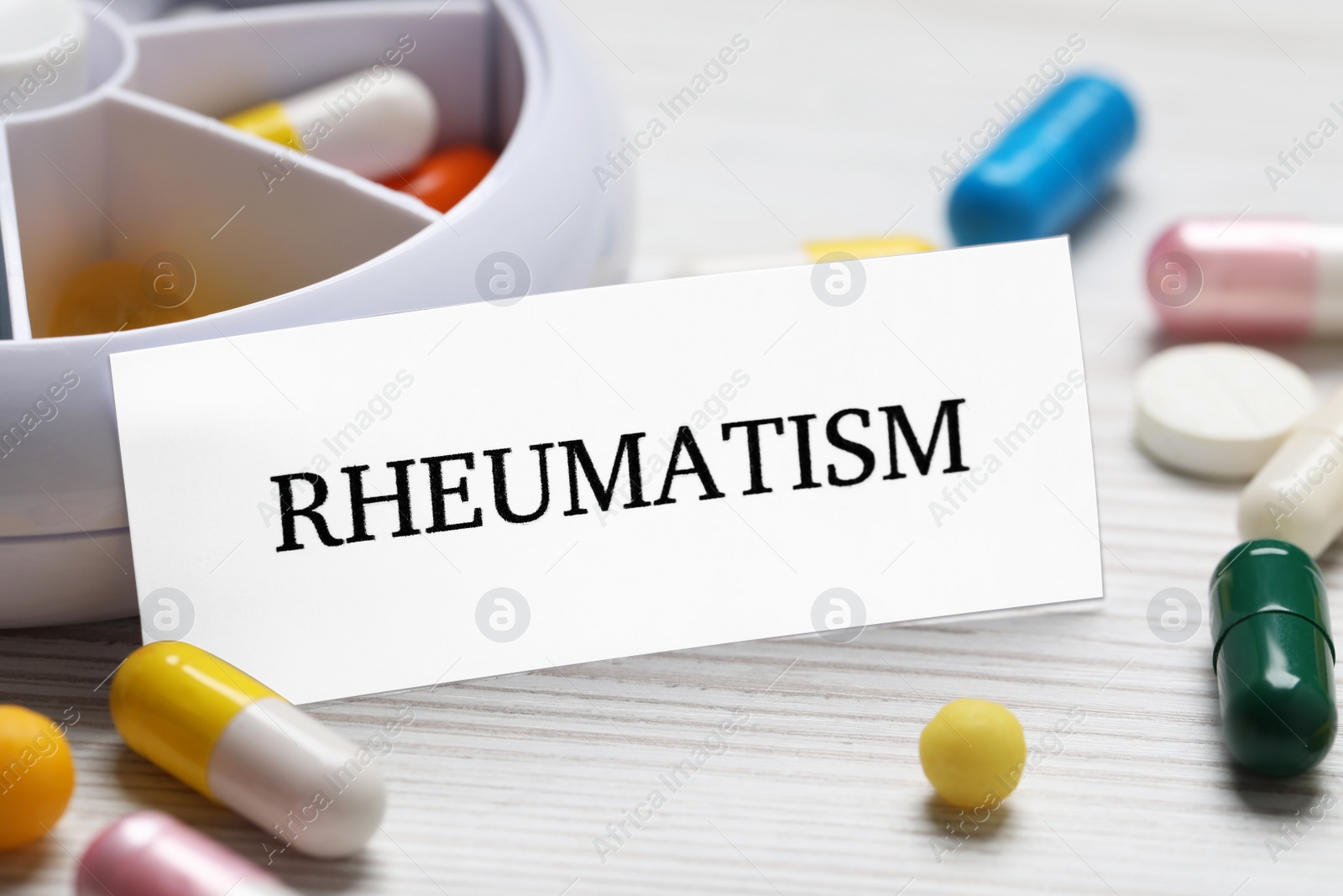 Photo of Card with word Rheumatism and pills on white wooden table, closeup