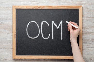 Woman writing abbreviation OCM (Organizational Change Management) on small blackboard with at white wooden table, top view