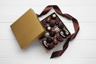 Photo of Open box of delicious chocolate candies and brown ribbon on white wooden table, flat lay