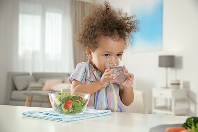 Photo of Cute African-American girl with glass of water and vegetable salad at table in living room