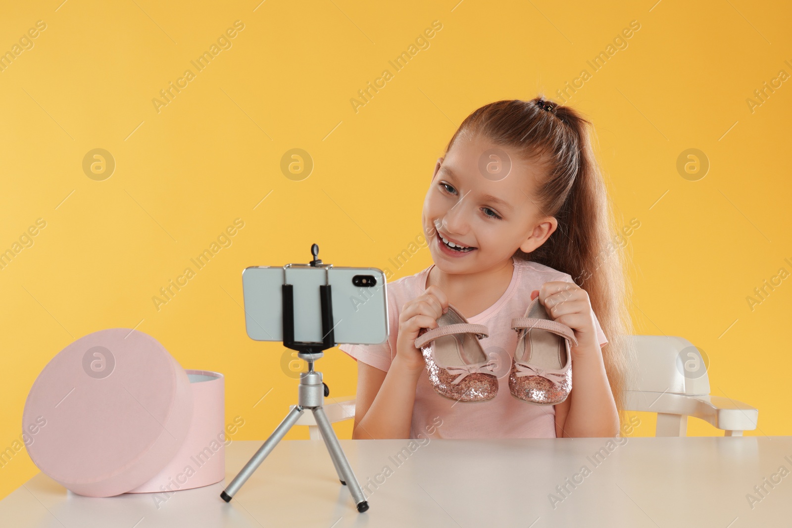 Photo of Cute little blogger with shoes recording video at table on yellow background