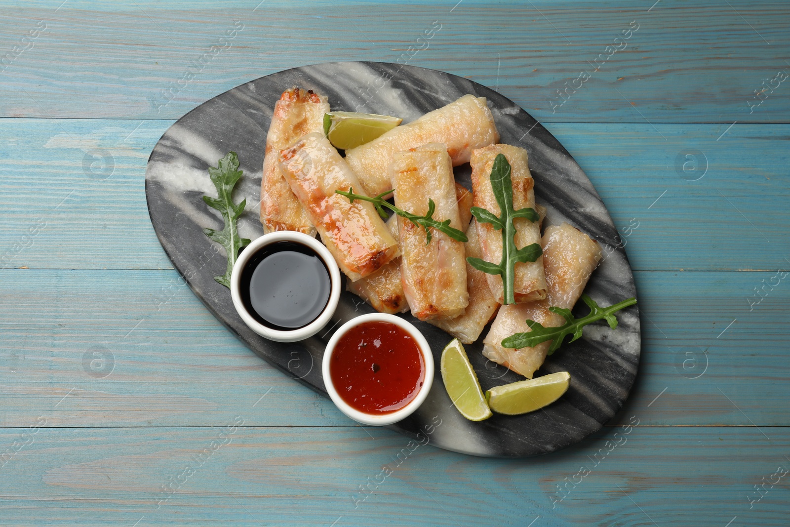 Photo of Tasty fried spring rolls, arugula, lime and sauces on light blue wooden table, top view