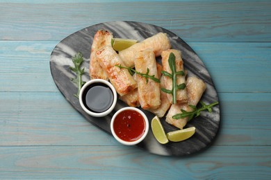 Photo of Tasty fried spring rolls, arugula, lime and sauces on light blue wooden table, top view