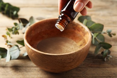 Photo of Woman dripping eucalyptus essential oil from bottle into bowl at wooden table, closeup