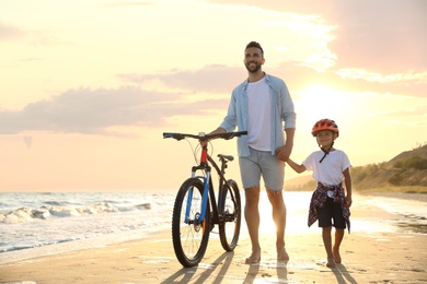 Photo of Happy father and son with bicycle on sandy beach near sea at sunset