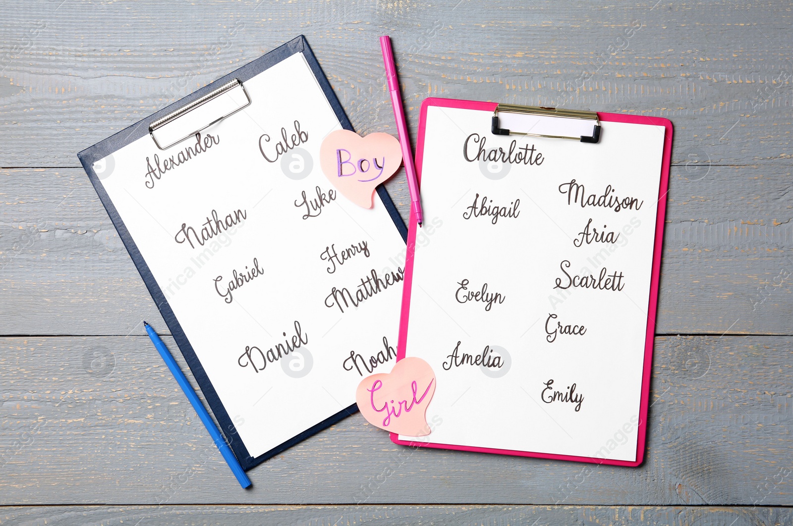 Photo of Clipboards with different baby names and felt tip pens on grey wooden table, flat lay