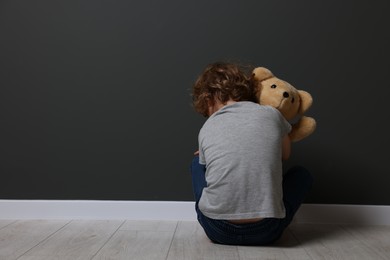 Photo of Child abuse. Upset boy with toy sitting on floor near grey wall, back view. Space for text