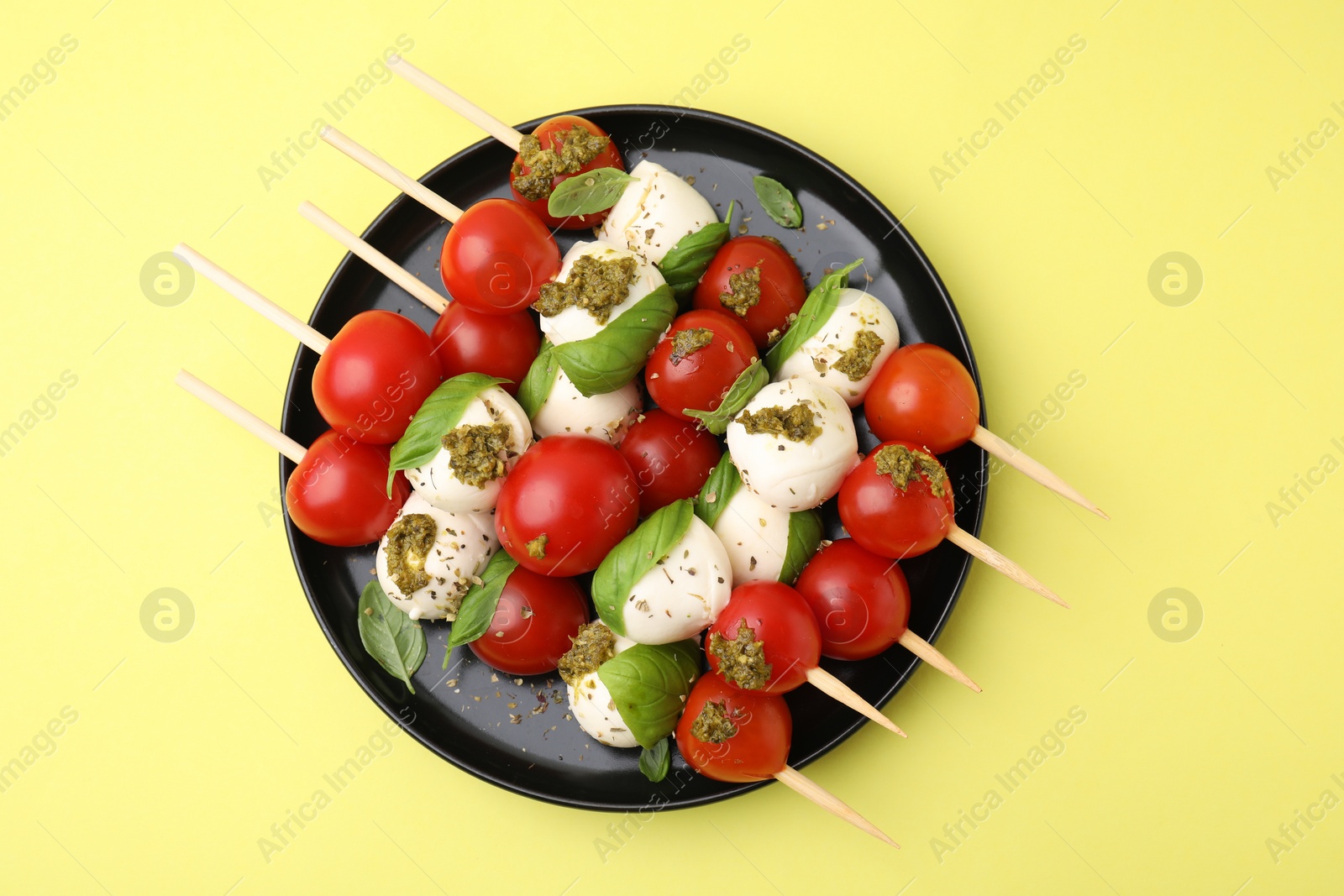 Photo of Caprese skewers with tomatoes, mozzarella balls, basil and pesto sauce on yellow background, top view