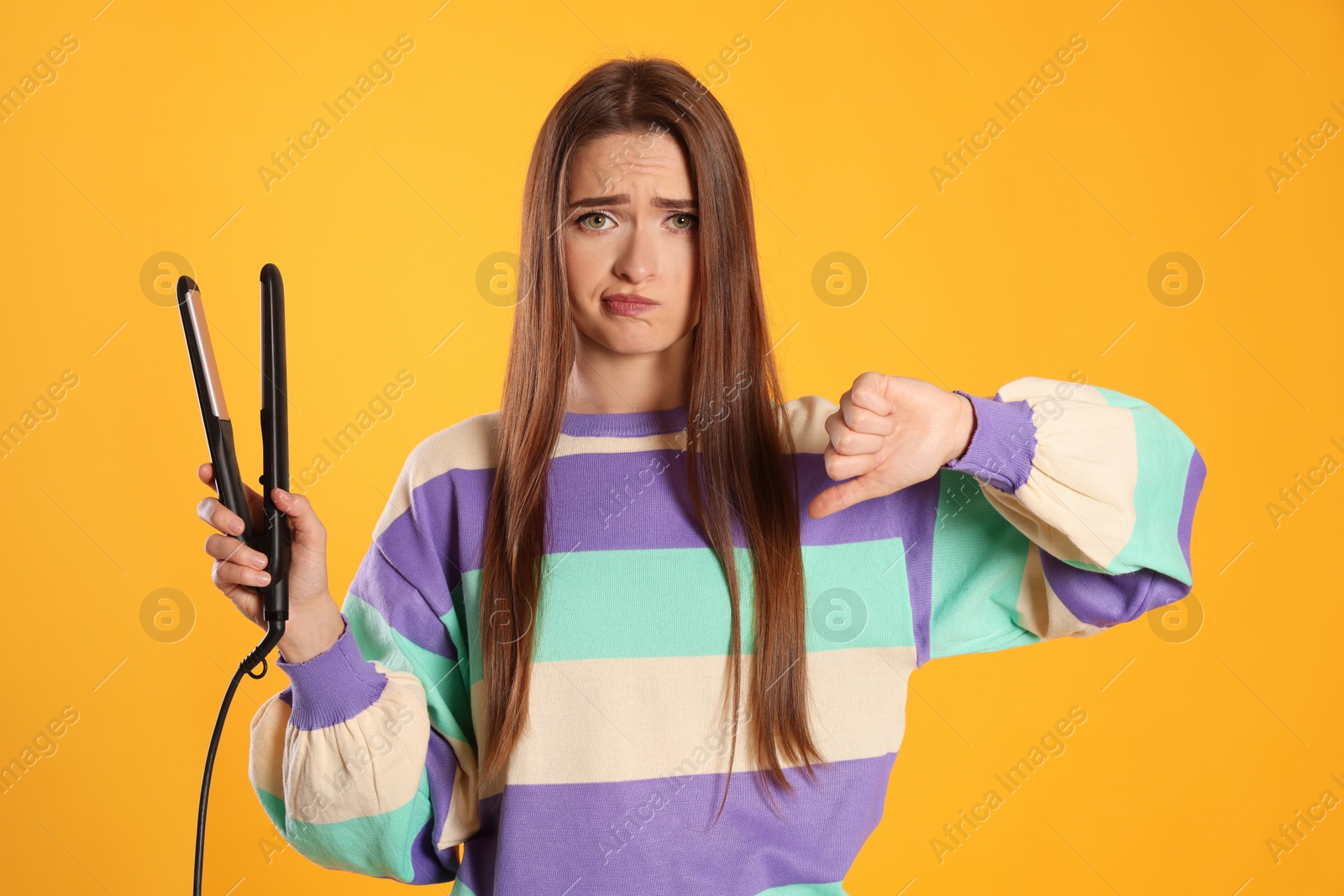 Photo of Upset young woman with flattening iron showing thumb down on yellow background. Hair damage