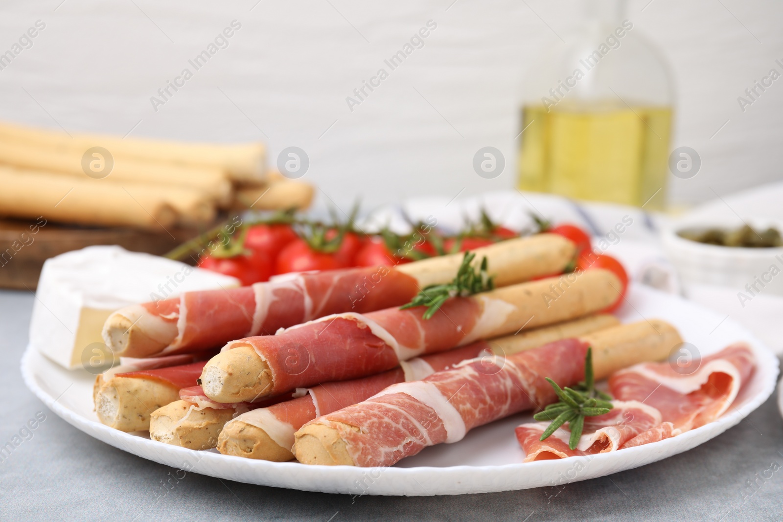 Photo of Plate of delicious grissini sticks with prosciutto, cheese and rosemary on light grey table, closeup