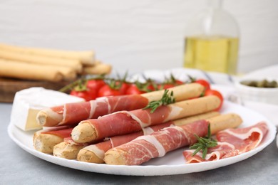 Photo of Plate of delicious grissini sticks with prosciutto, cheese and rosemary on light grey table, closeup