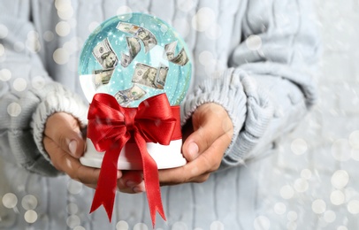 Image of Woman holding snow globe with dollar banknotes, closeup. Bokeh effect 