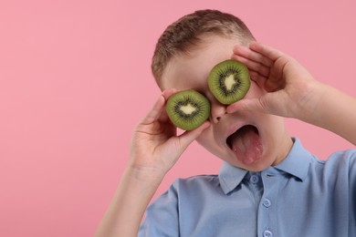 Photo of Emotional boy covering eyes with halves of fresh kiwi and showing tongue on pink background, space for text