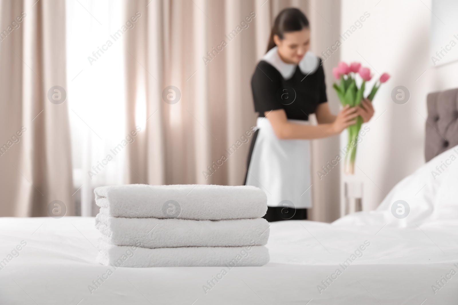 Photo of Young maid with flowers in hotel room, focus on stack of towels