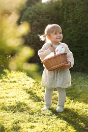 Photo of Happy little girl holding wicker basket with cute rabbit outdoors on sunny day