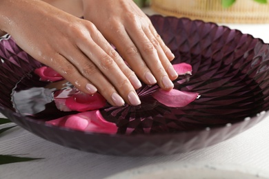 Photo of Woman soaking her hands in bowl with water and petals on table, closeup. Spa treatment