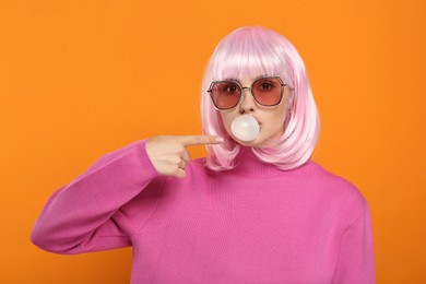 Photo of Beautiful woman in sunglasses blowing bubble gum and pointing at herself on orange background