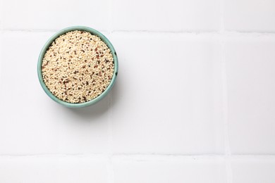 Photo of Raw quinoa seeds in bowl on white tiled table, top view. Space for text