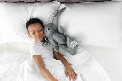 Photo of Cute little African-American boy with toy rabbit sleeping in bed, top view