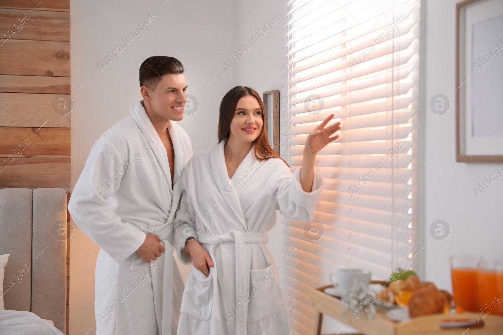 Photo of Happy couple wearing bathrobes near window in bedroom