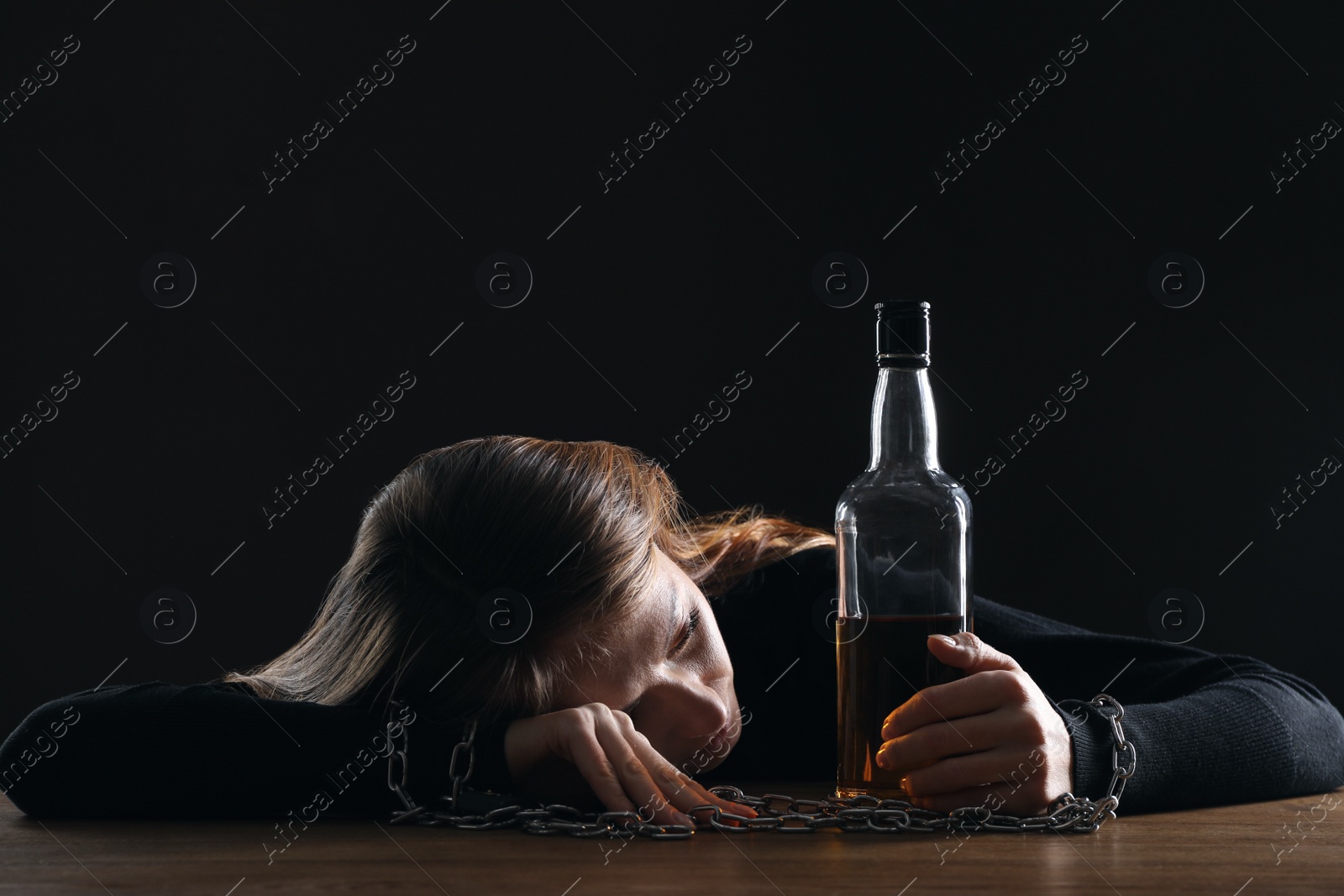 Photo of Alcohol addiction. Woman chained with bottle of whiskey at wooden table against black background