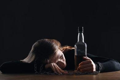 Alcohol addiction. Woman chained with bottle of whiskey at wooden table against black background