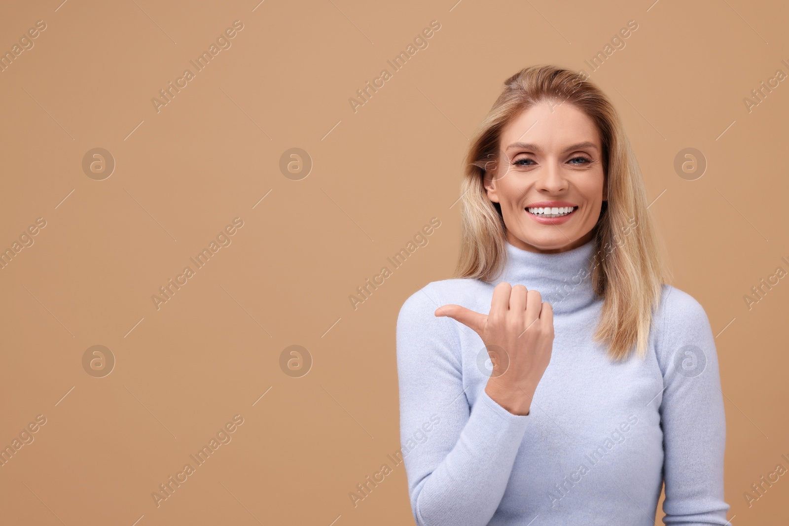 Photo of Portrait of smiling middle aged woman with blonde hair pointing at something on beige background. Space for text