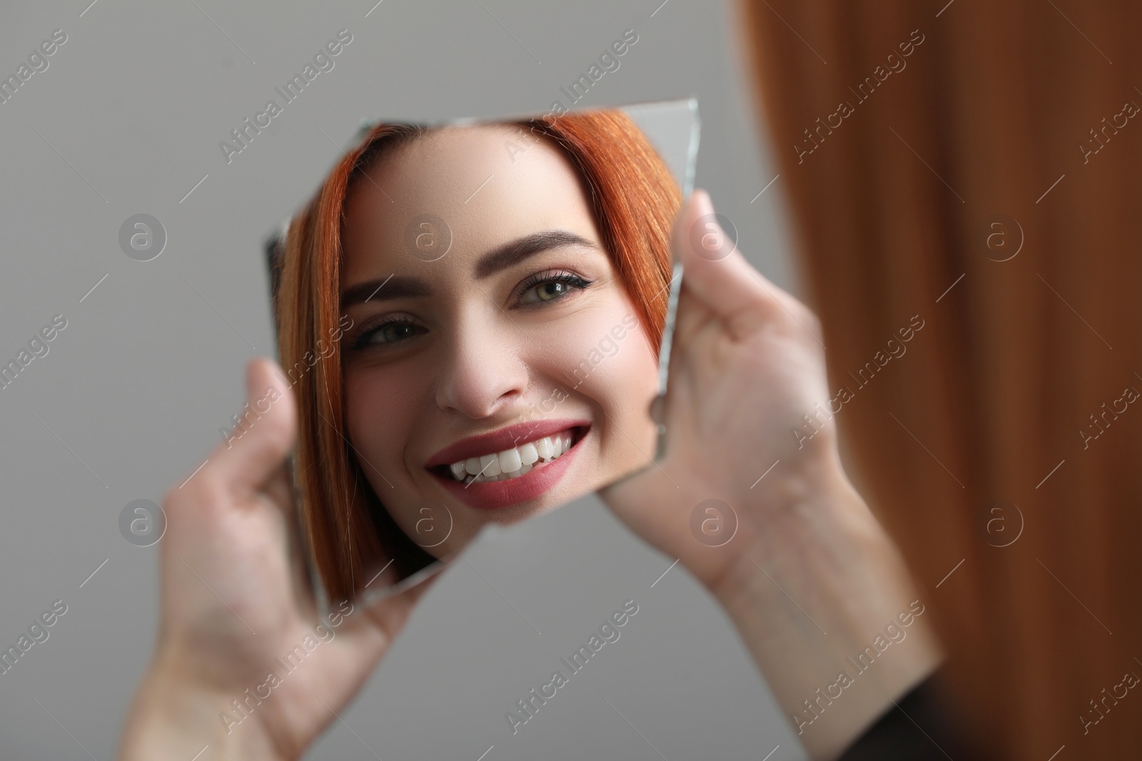 Photo of Smiling young woman looking at herself in broken mirror on light grey background, closeup