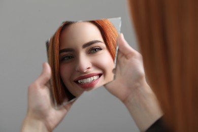 Smiling young woman looking at herself in broken mirror on light grey background, closeup