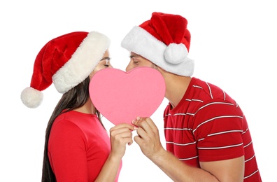 Photo of Young happy couple with Santa hats hiding and kissing behind pink heart on white background. Christmas celebration