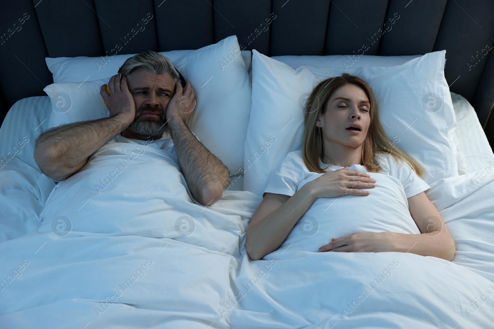 Photo of Irritated man with headphones lying near his snoring wife in bed at home