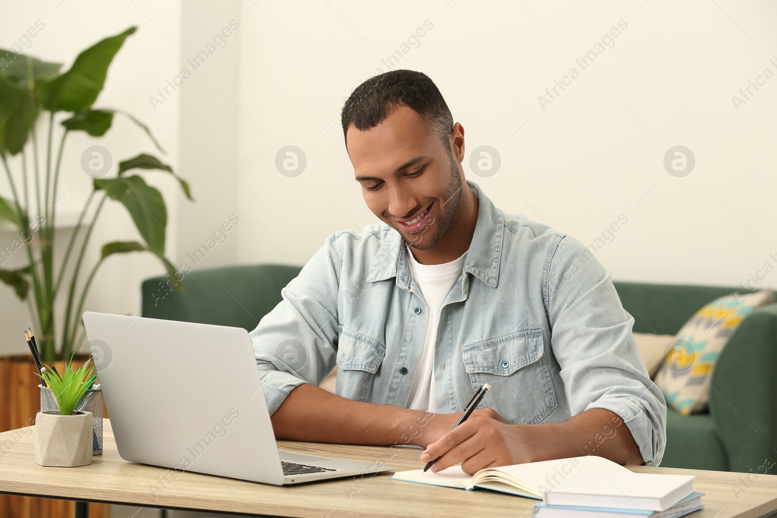 Photo of African American man writing in notepad near laptop at wooden table in room