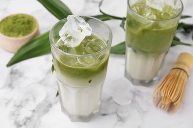 Photo of Glasses of tasty iced matcha latte, bamboo whisk and leaves on white marble table, closeup