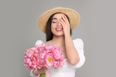 Photo of Beautiful young woman in straw hat with bouquet of pink peonies against grey background