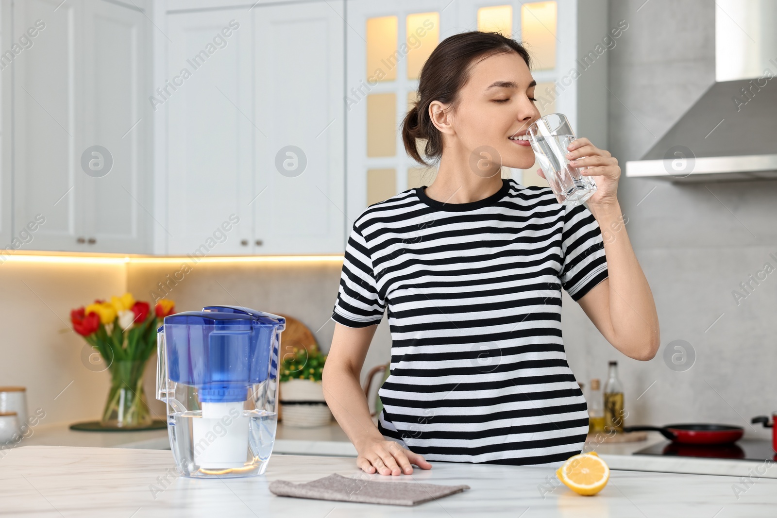 Photo of Woman drinking water and filter jug in kitchen