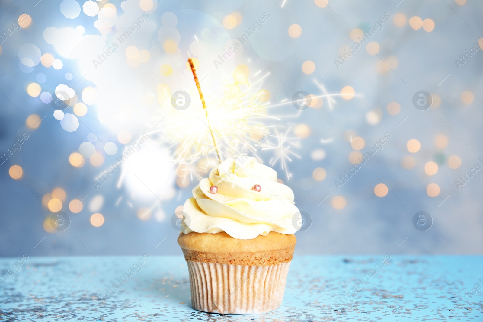 Image of Delicious birthday cupcake with sparkler on blue table against blurred lights