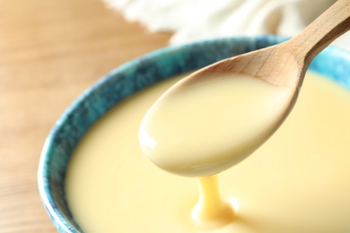 Photo of Spoon of pouring condensed milk over bowl on table, closeup with space for text. Dairy products