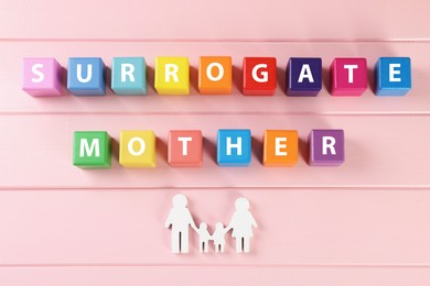 Phrase Surrogate Mother of colorful cubes and paper family cutout on pink wooden table, flat lay