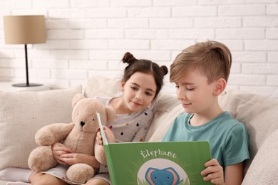 Photo of Little boy reading book to girl with toy bear on sofa at home