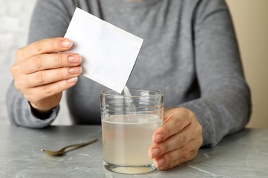 Photo of Woman pouring powder from medicine sachet into glass of water on grey marble table, closeup