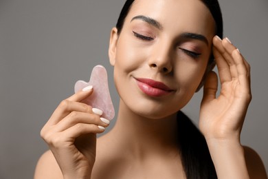 Beautiful young woman doing facial massage with gua sha tool on grey background, closeup