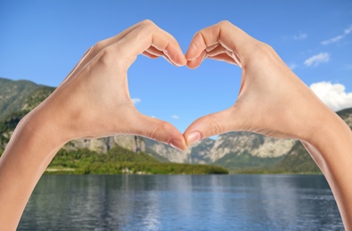 Image of Woman making heart with hands near river and mountains on sunny day, closeup