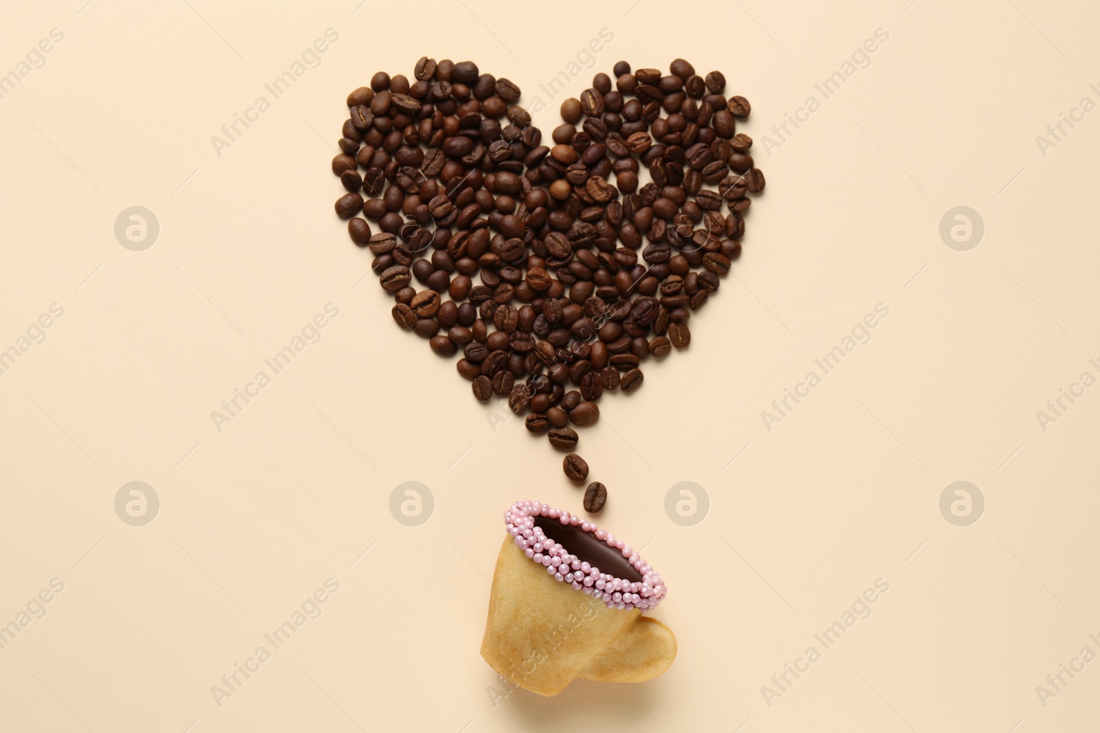 Photo of Coffee beans spilled from edible biscuit cup in shape of heart on beige background, top view