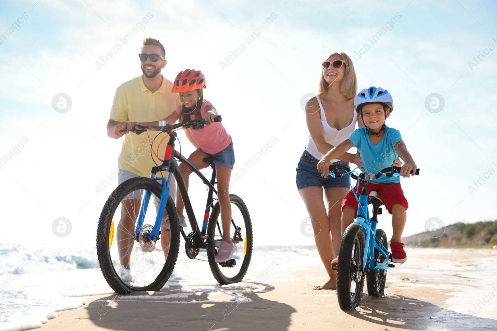 Photo of Happy parents teaching children to ride bicycles on sandy beach near sea