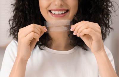 Photo of Young woman holding teeth whitening strips indoors, closeup