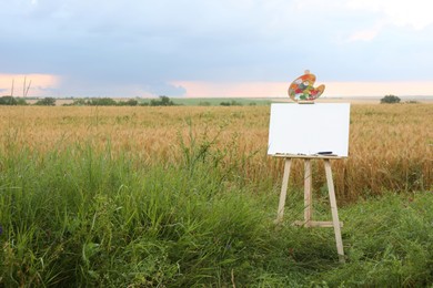 Photo of Wooden easel with blank canvas and painting equipment in field. Space for text