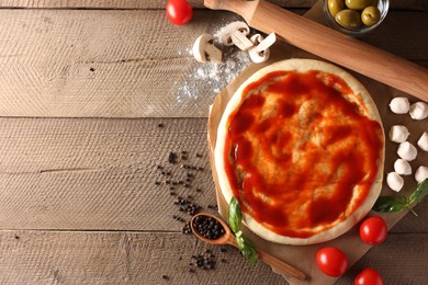 Pizza dough smeared with tomato sauce, products and rolling pin on wooden table, flat lay. Space for text