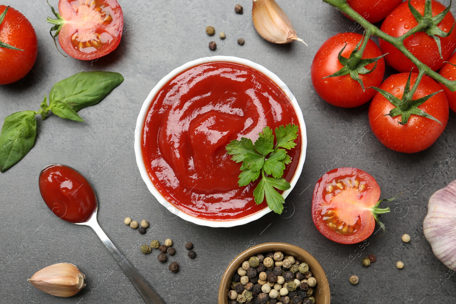 Photo of Delicious tomato ketchup in bowl, spices and products on grey textured table, flat lay