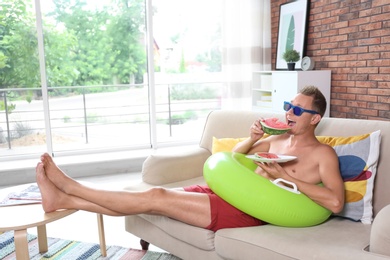 Photo of Shirtless man with inflatable ring eating watermelon on sofa at home